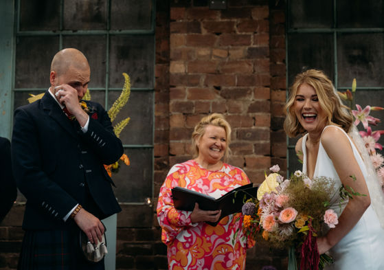 Celebrant in bright clothing smiles while bride laughs and groom wipes tears